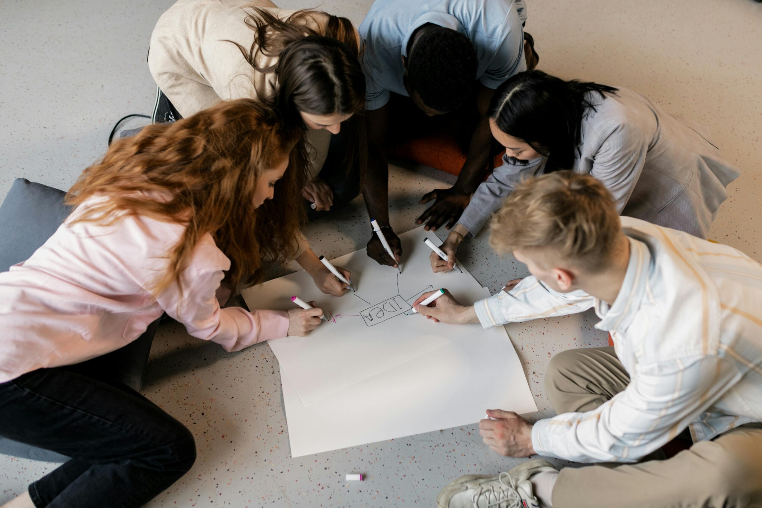 A multicultural team collaborates on an idea using markers and paper on the floor indoors.